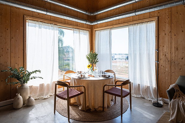 mesa redonda junto a ventana con cortinas blancas, paredes de madera en una casa pasiva
